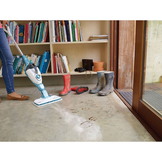 BLACKDECKER FSMH1321J steam mop cleaning muddy footprints on concrete floor near boots and bookshelf.