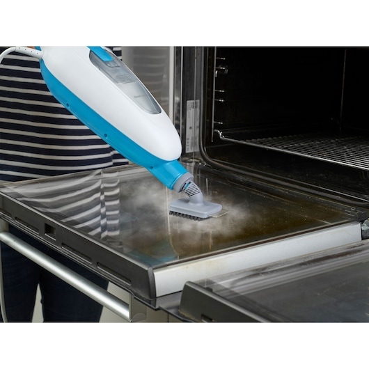 A steam cleaner with a blue and white body is being used to clean the inside door of an oven, emitting steam as it removes dirt.