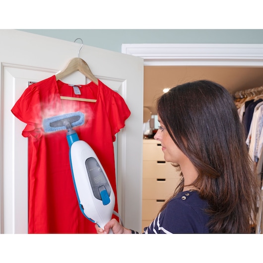 A person uses a BLACK+DECKER handheld steam cleaner to steam a red blouse hanging on a door in a closet area. The steam appliance is visibly emitting steam onto the fabric.
