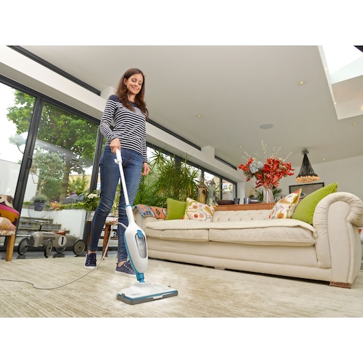 A person using a BLACK+DECKER steam mop to clean a carpet in a modern living room with a beige sofa, colorful cushions, and large windows.