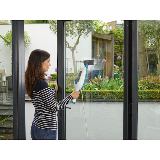 A person with blurred face is using a BLACK+DECKER steam cleaner to clean a glass door in a modern home setting. The steam cleaner is white and blue, and the glass door looks out onto a garden with green plants and purple flowers.
