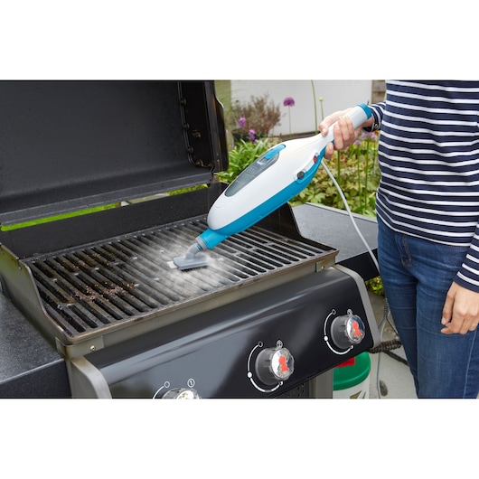 A person is using a handheld steam cleaner to clean the grill of an outdoor barbecue.