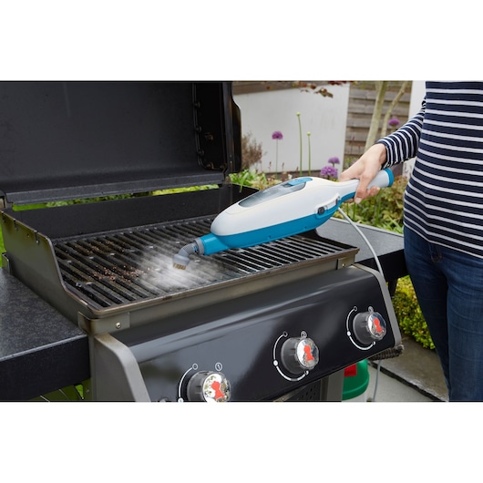 A person is using a BLACK+DECKER steam cleaning device to clean the grill grates of an outdoor barbecue, emitting steam as it removes dirt.