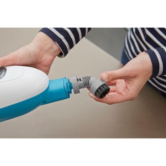 A person is attaching a small brush accessory to a blue and white cleaning device, possibly a steam cleaner.