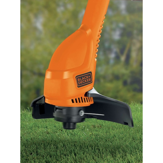Close-up of a BLACK+DECKER electric grass trimmer being used on a lawn, featuring an orange body and black cutting head, with greenery in the background.