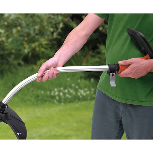 A person is holding a BLACK+DECKER grass trimmer with both hands outdoors, adjusting the handle. The background shows a garden with green grass and plants.