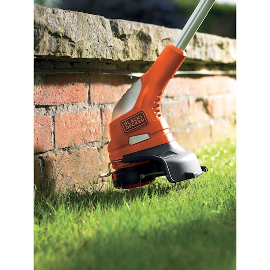 A close-up view of a BLACK+DECKER cordless grass trimmer being used to trim the grass near a brick wall.