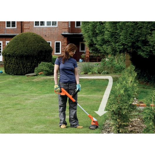 Person wearing gloves and work clothes using a BLACK+DECKER cordless grass trimmer to tidy the lawn edge near a hedge. The scene is outdoors with a red brick house and garden visible in the background.