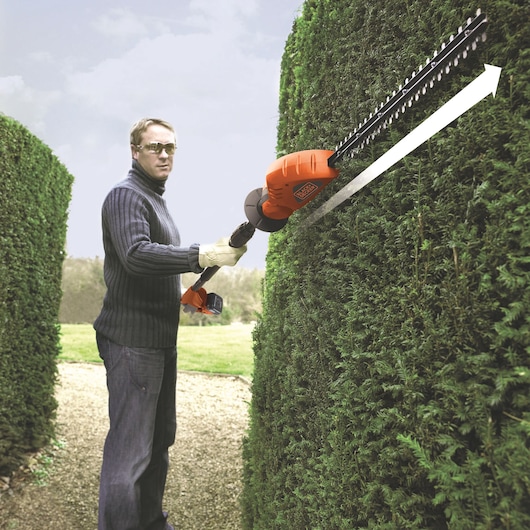 BLACKDECKER GTC1843L20 hedge trimmer used outdoors on tall green hedge, side angle.
