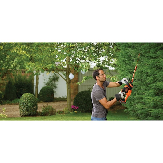 Person trimming tall hedge with BLACKDECKER cordless hedge trimmer GTC18452PC in a garden.