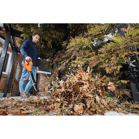 A person uses an orange BLACK+DECKER cordless leaf blower to clear dry leaves from a garden patio.