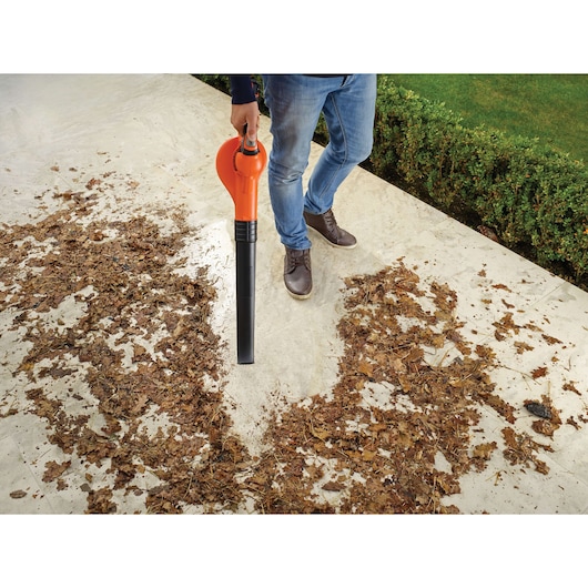 A person is using an orange and black leaf blower to clear dried leaves from a paved outdoor area. The leaf blower is held in one hand and is removing leaves, creating a clean path on the ground. The person is wearing blue jeans and brown shoes. There is a green hedge and lawn in the background.