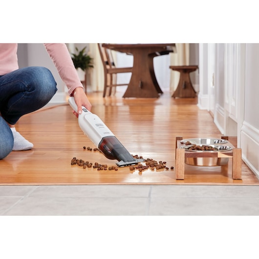 BLACKDECKER HLVC315B11 handheld vacuum cleaning pet food from wooden floor near dog bowl, side angle.