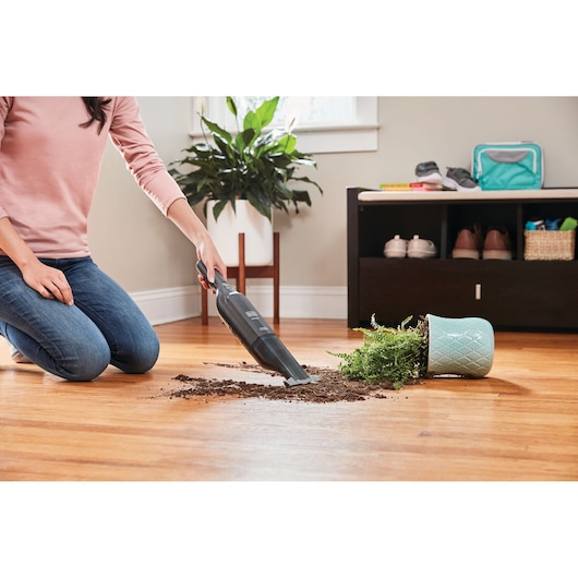 BLACKDECKER HLVC320B11 handheld vacuum cleaning spilled soil near tipped plant on hardwood floor.
