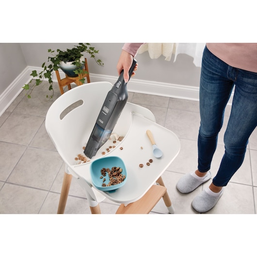 BLACKDECKER HLVC320J11 handheld vacuum cleaning cereal from a high chair tray in a kitchen.