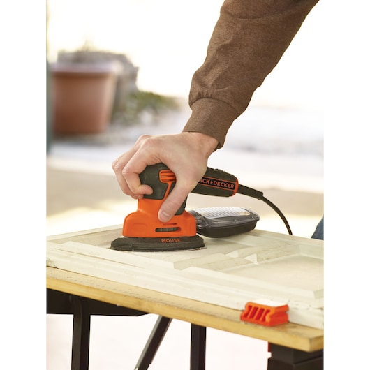 Close-up of a person using a BLACK+DECKER Mouse sander on a wooden surface. The sander is orange and black, and the person is working on a light-colored piece of wood placed on a table.