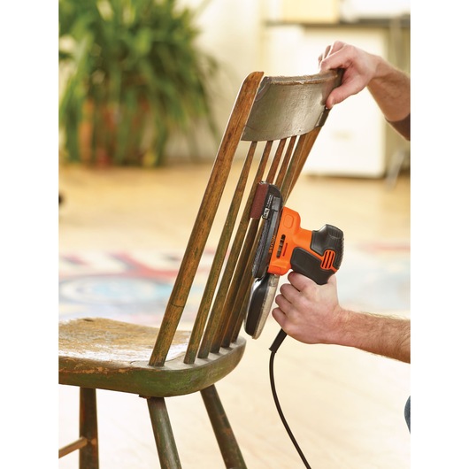 A person is using a BLACK+DECKER electric sander to sand the backrest of a wooden chair.