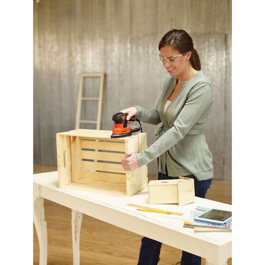 A person using a BLACK+DECKER KA2000AT sander to smooth a wooden crate placed on a white table, with additional wood pieces, a ruler, pencil, and a tablet nearby. The person's face is blurred, and the workspace has a corrugated metal wall and a wooden floor.