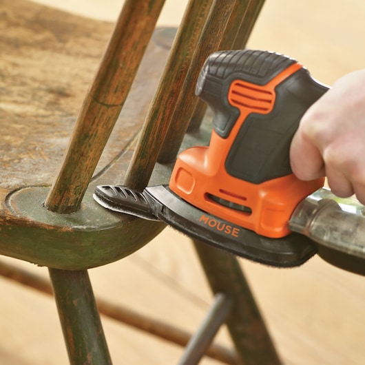 A close-up of a BLACK+DECKER mouse sander being used to sand the corner of a wooden chair, focusing on the tool and the surface being worked.
