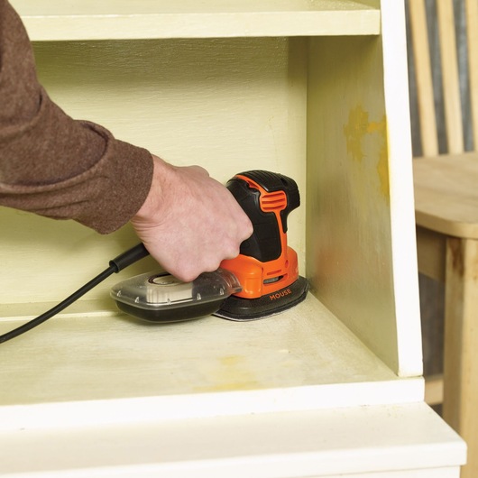 A person using an orange and black BLACK+DECKER electric sander to smooth the inside shelf of a wooden cabinet.