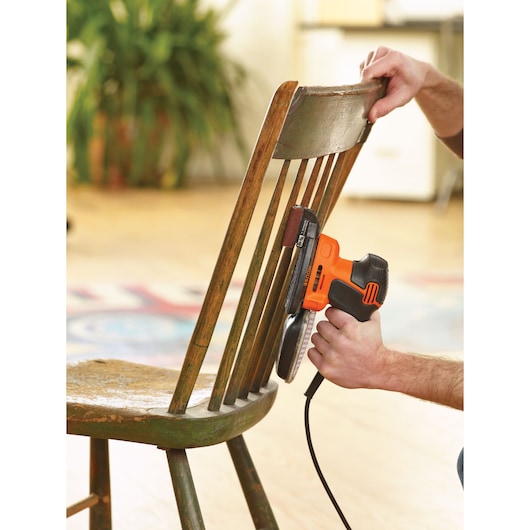 A person uses a BLACK+DECKER KA2000KA10 electric sander to sand the backrest of a wooden chair indoors.