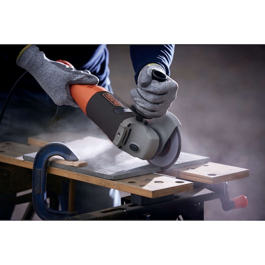 A close-up view of a person using a BLACK+DECKER angle grinder to cut a tile on a workbench. The person is wearing protective gloves and the workspace is secured with a clamp.