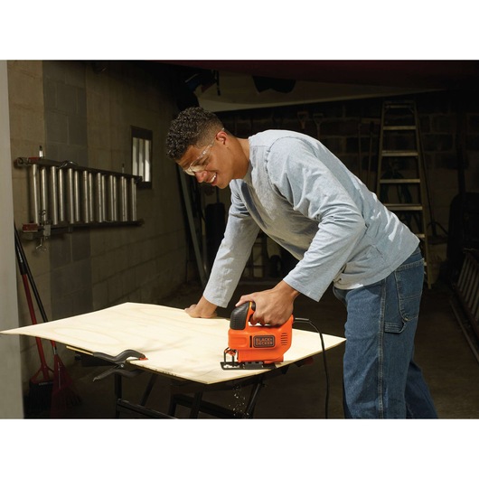A person using a BLACK+DECKER KS701E jigsaw to cut a piece of wood on a workbench in a garage or workshop. The individual is wearing a light gray long-sleeve shirt and jeans. Various tools and equipment, including a ladder and pipes, are visible in the background.