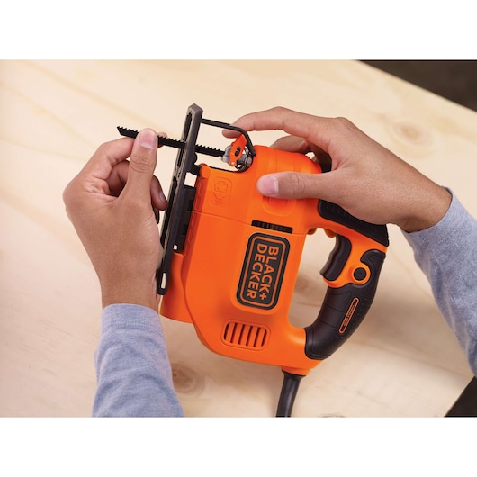 A person is installing a saw blade into an orange and black BLACK+DECKER jigsaw on a light wooden surface.