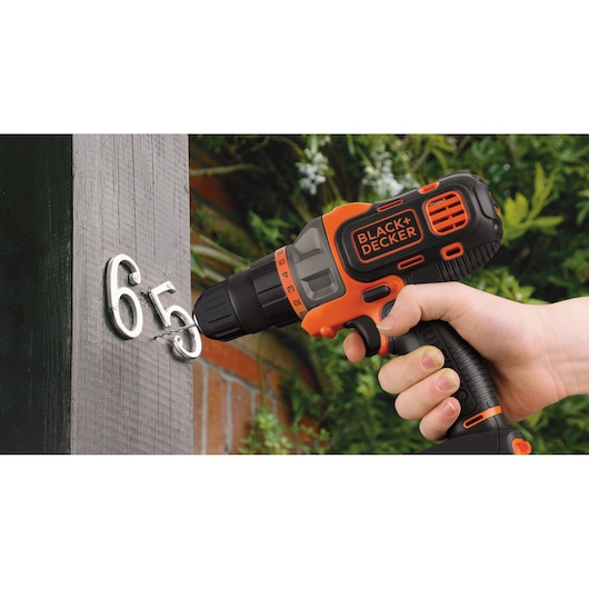 BLACK+DECKER MT218K cordless drill in use, drilling metal numbers into outdoor wood post.