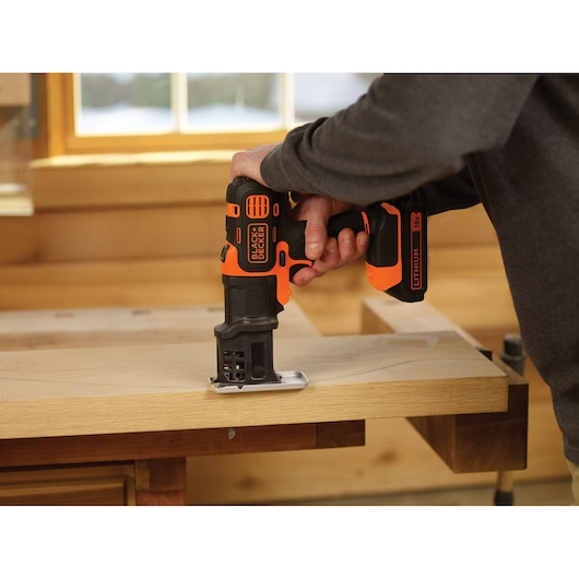 A person is using a BLACK+DECKER cordless jigsaw to cut a piece of wood on a workbench in a woodworking workshop.