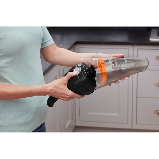 Person emptying BLACKDECKER handheld vacuum NVC115BJL with transparent dust container in a kitchen.