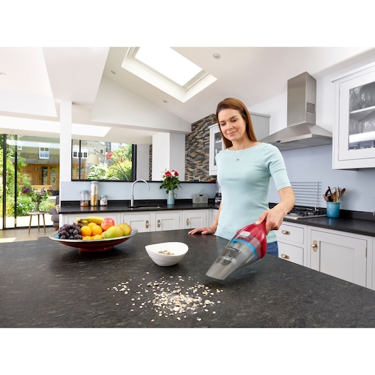 BLACKDECKER NVC115WAR red handheld vacuum cleaning cereal on a kitchen counter near a bowl and fruit plate.