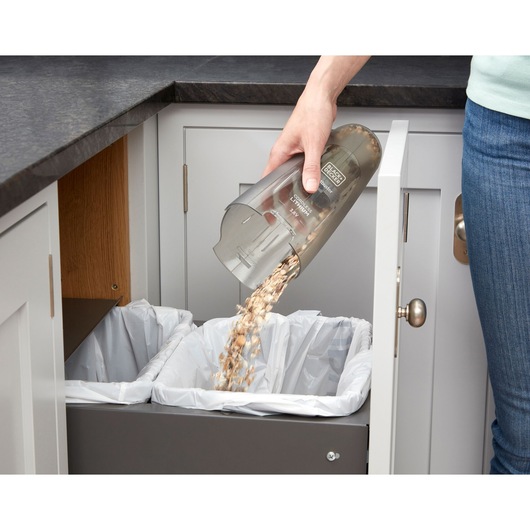 BLACK+DECKER NVC115W handheld vacuum canister being emptied into a kitchen trash bin.