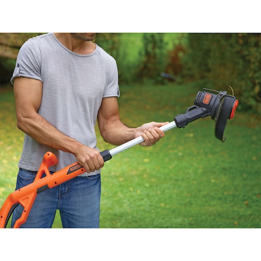 A person using a BLACK+DECKER grass trimmer outdoors, holding the orange and black tool above green grass.