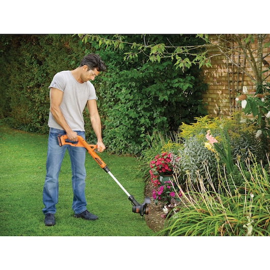 A person is using an orange BLACK+DECKER cordless grass trimmer (model ST1823) to edge the lawn near a flower bed in a garden.