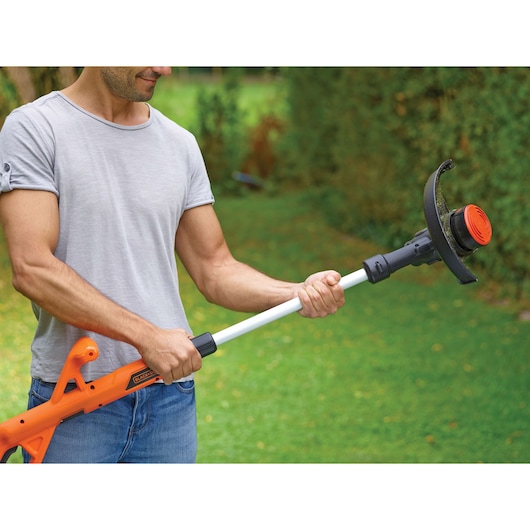 A person holding a BLACK+DECKER ST1823 orange and black grass trimmer in a garden, with the face blurred for privacy.