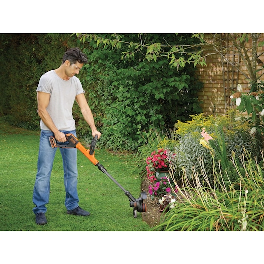 A person using a BLACK+DECKER cordless grass trimmer to edge a garden lawn near flower beds. The person is outdoors, standing on grass, trimming the edge of a garden with various colorful plants and flowers. Their face is blurred.
