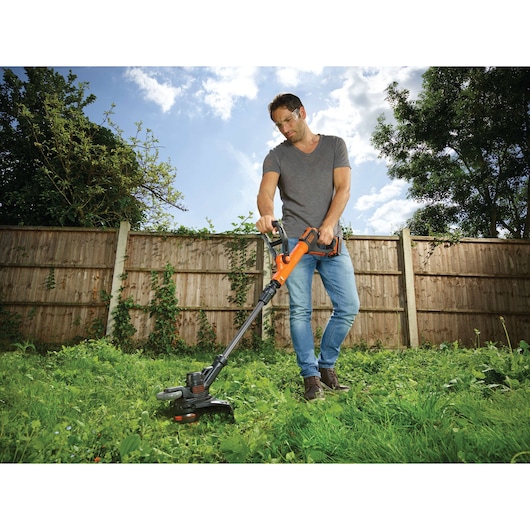 A person using an orange and black BLACK+DECKER cordless grass trimmer on a lawn in a backyard, with a wooden fence and trees in the background.
